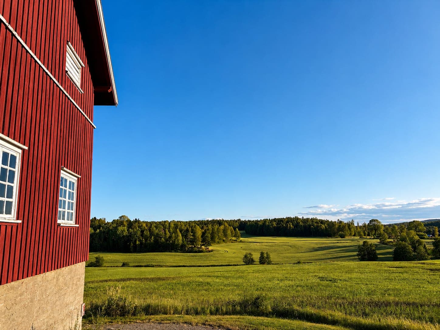 Rød låve med hvite vinduer, med grønne jorder og skog bakenfor under blå himmel